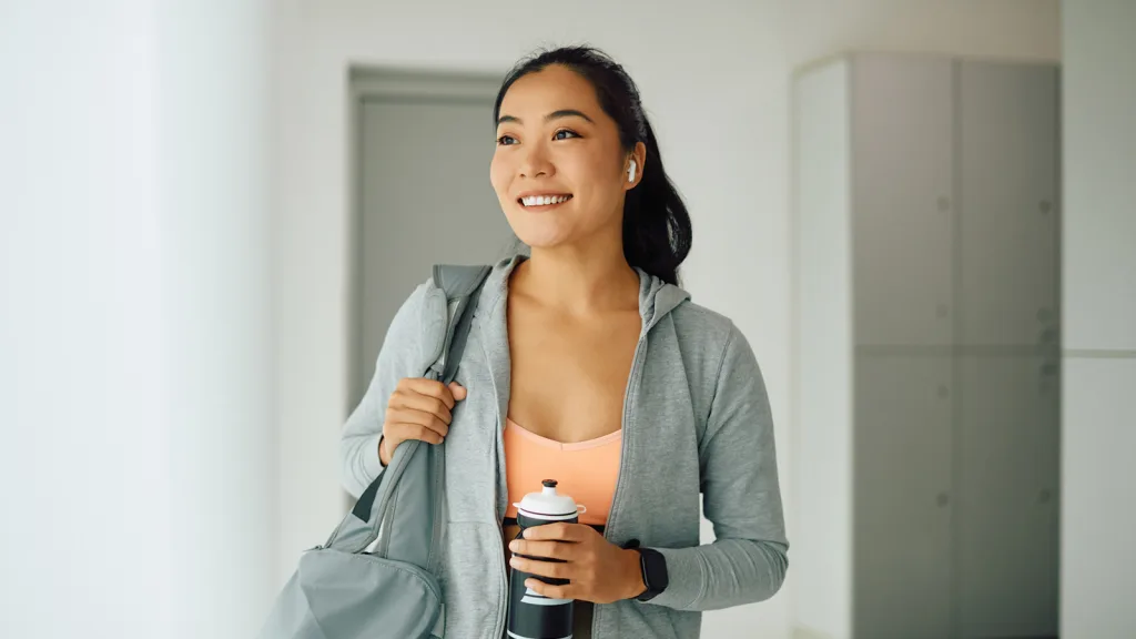 Woman smiling in an empty locker room, she is wearing an athletic top with a sweater unzipped where you can see her olive-green athletic leggings. She is wearing an apple watch and holding her water bottle with her left arm while her right arm braces her gym bag on her right shoulder. She isn't looking at the camera but possible out a window. New pet-friendly apartment homes for rent with private fenced backyards in Arizona, Texas, Florida, and North Carolina with pool, pickleball, dog park, gym, clubhouse and gated community. *No breed or weight restrictions. Yardly