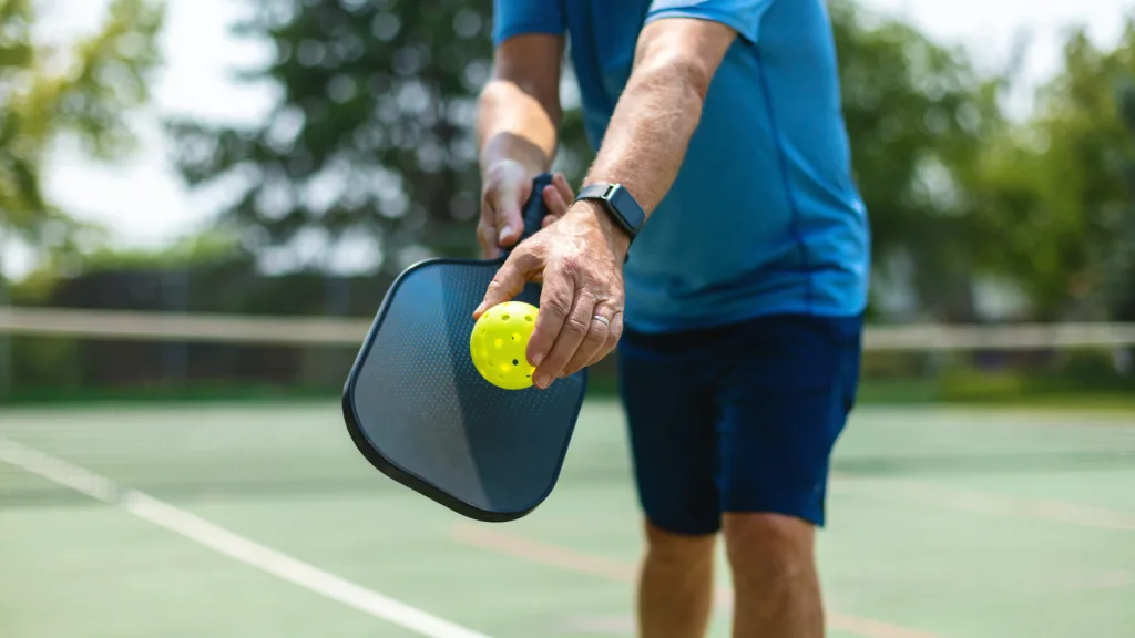 Man standing in a serving stance, holding a pickleball and racket. He is in the background wearing navy blue athletic shorts and a blue athletic top. He has a smart watch on his wrist and greenery and foliage behind him. New pet-friendly apartment homes for rent with private fenced backyards in Arizona, Texas, Florida, and North Carolina with pool, pickleball, dog park, gym, clubhouse and gated community. *No breed or weight restrictions. Yardly