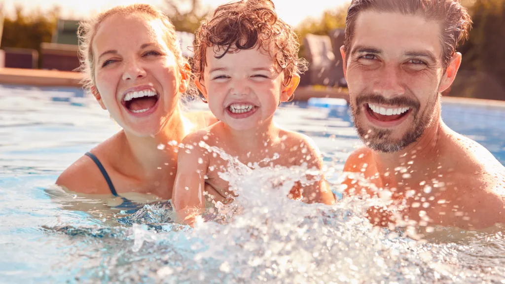 Family splashing and laughing in the pool. Mom on the left hair slicked back laughing holding toddler son smiling and splashing in the water with dad on the right has a laughing smile with a short shaggy beard also holding him all looking at the camera in a candid moment in the pool. New pet-friendly apartment homes for rent with private fenced backyards in Arizona, Texas, Florida, and North Carolina with pool, pickleball, dog park, gym, clubhouse and gated community. *No breed or weight restrictions. Yardly